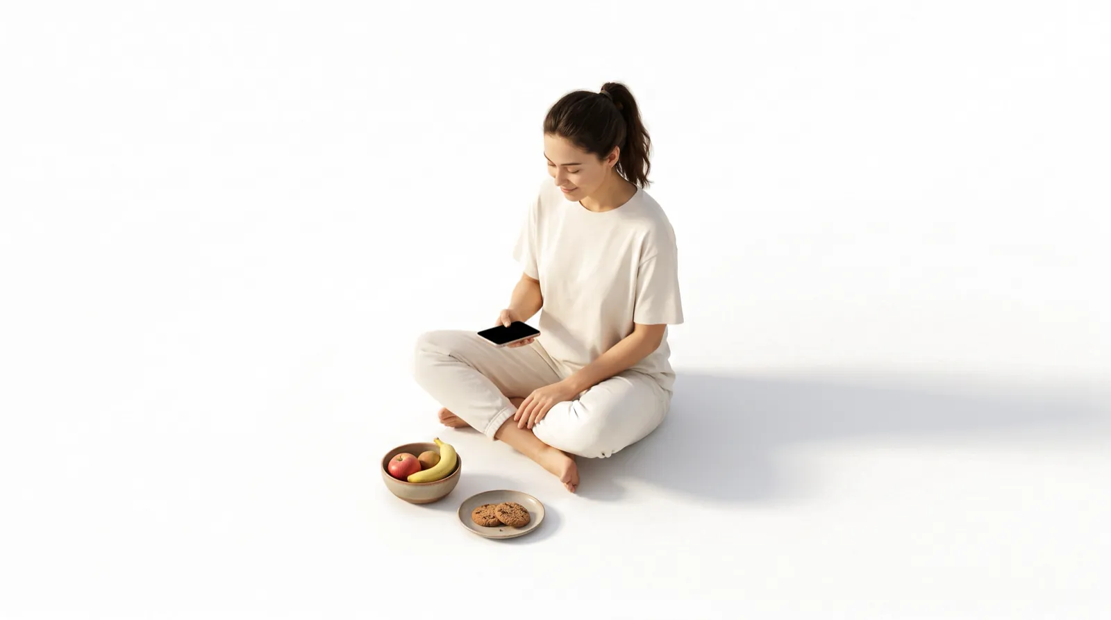 A woman sitting cross-legged looking at her phone, with fruit and a few cookies beside her